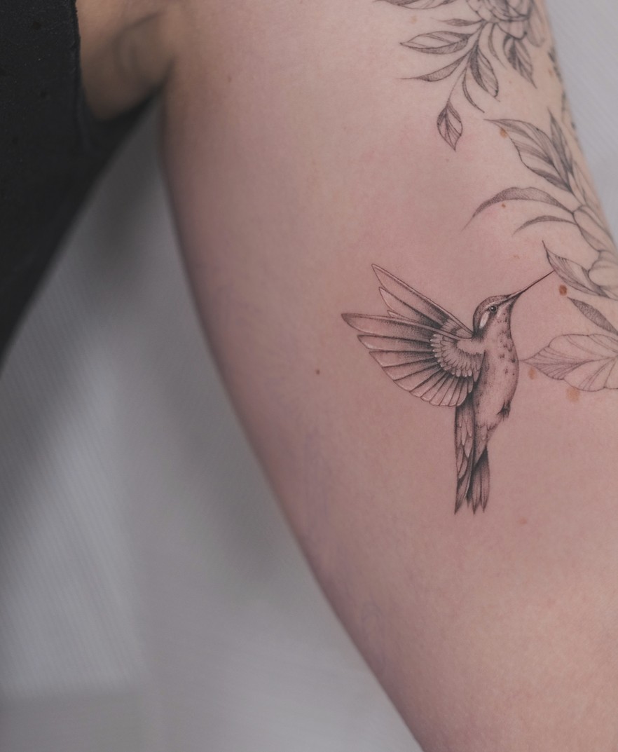 Fineline tattoo van een kolibrie tussen fijne bloemen op de arm