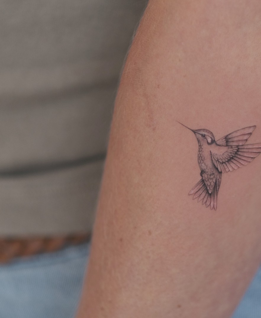 Kleine fineline tattoo van een kolibrie op de bovenarm